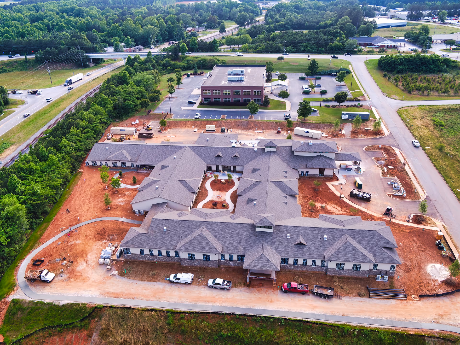 Aerial view of a large building under construction with a U-shaped layout, surrounded by red dirt and construction vehicles. The building is located near a road with green trees and other buildings in the background.