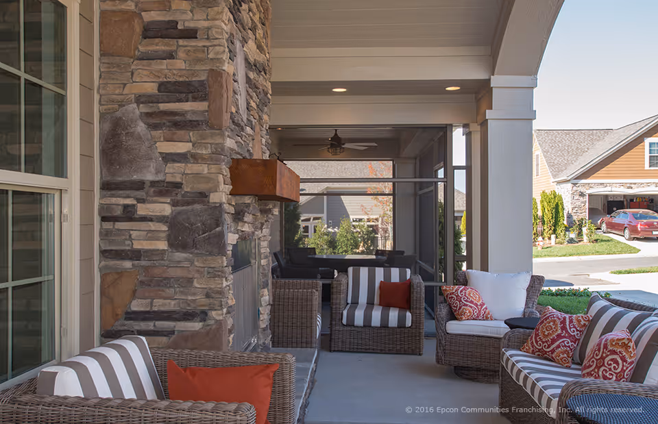Covered outdoor patio area with wicker furniture including chairs and a sofa with striped and patterned cushions. There is a stone fireplace on the left side and a ceiling fan above. The patio overlooks a residential neighborhood with houses and a red car parked in a driveway.