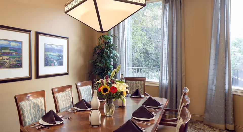 A formal dining room with a wooden table set for six people. Each place setting has a white plate with a folded dark napkin, silverware, and a glass. A vase with a colorful flower arrangement is centered on the table. The room has two large windows with light blue curtains, two framed landscape paintings on the wall, and a large hanging light fixture above the table. There is a green potted plant in the corner near the windows.