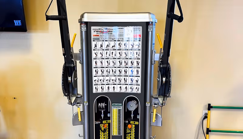 Multi-functional cable weight machine with exercise chart and handles in an indoor fitness area.