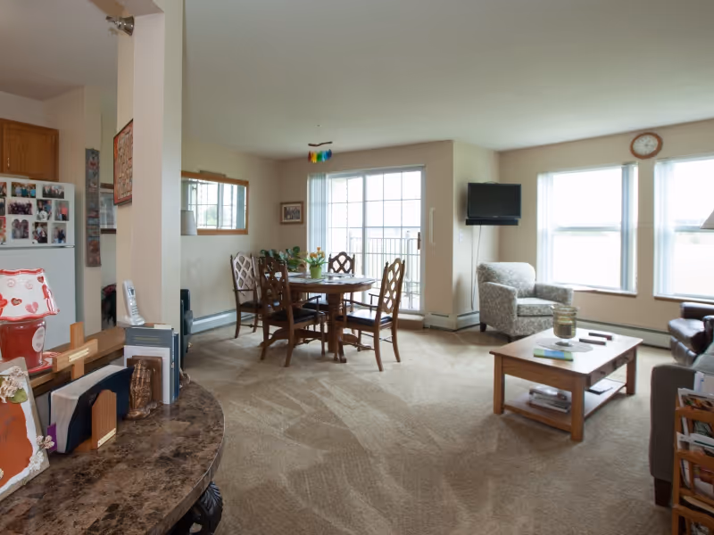 Carpeted open living and dining area with a round wooden dining table by sliding glass doors, armchairs, a coffee table, and a wall-mounted TV.