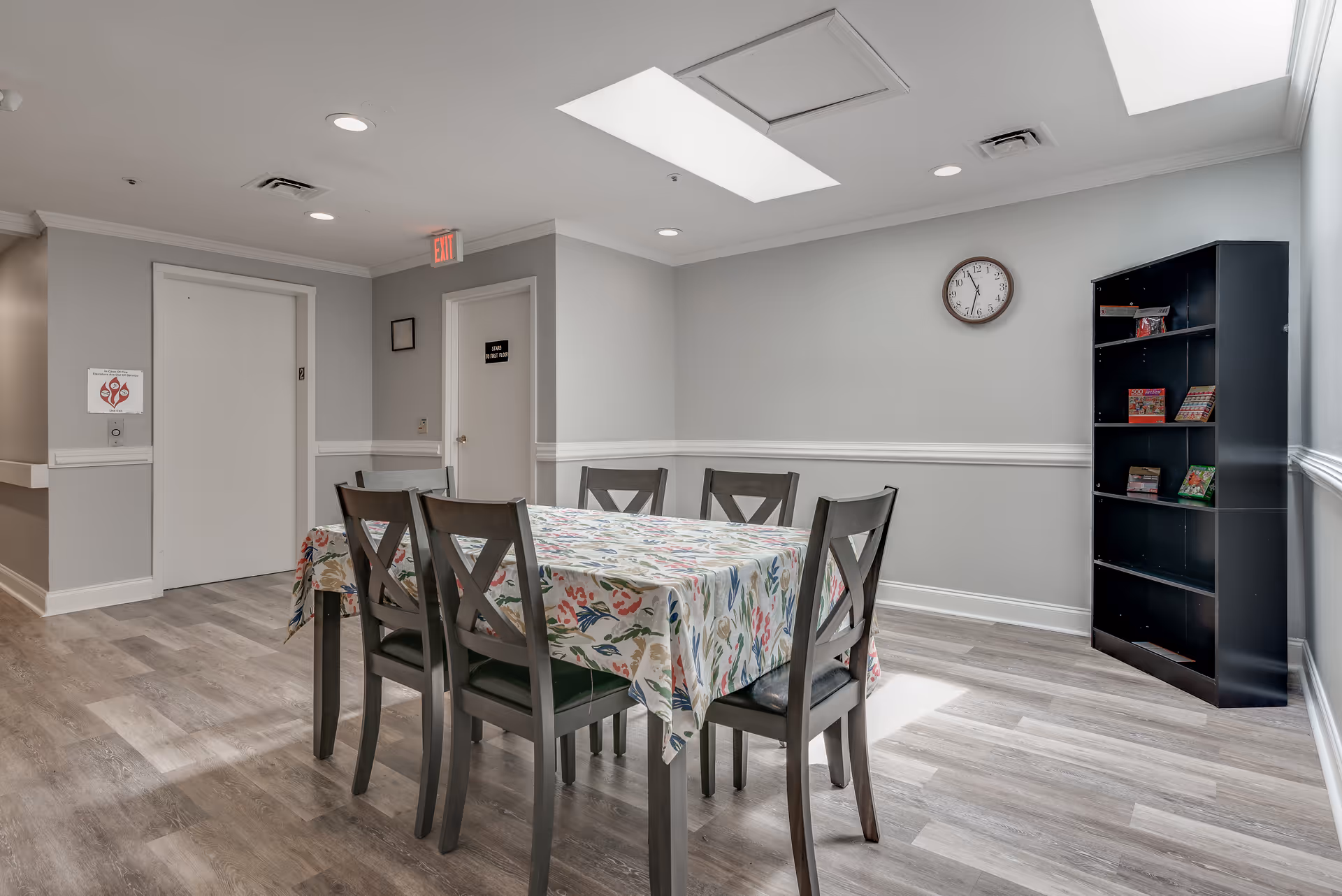 Small communal dining room with a floral-covered table and six chairs, a wall clock, and a black bookshelf.