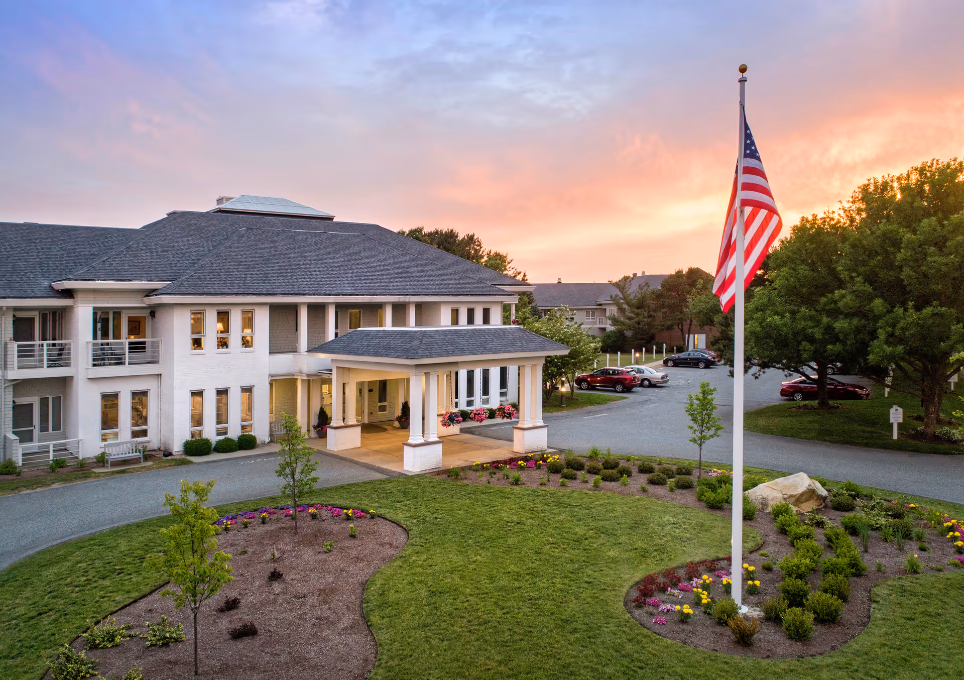Exterior view of a senior living facility building at sunset with a landscaped garden and an American flag on a flagpole in the foreground. Several cars are parked in the parking area near the building.