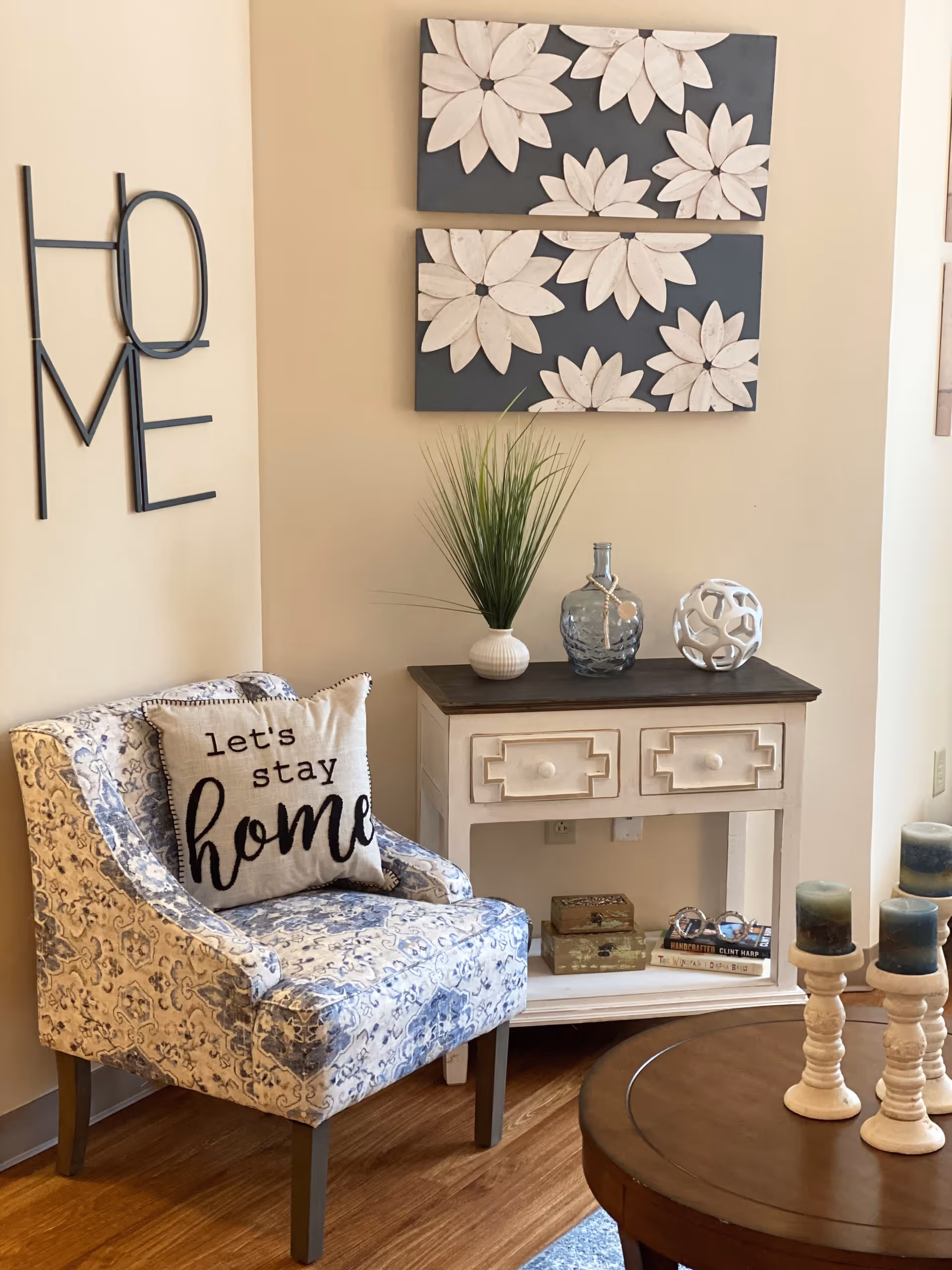 A cozy corner of a living room featuring a patterned armchair with a pillow that says 'let's stay home', a small white wooden console table with decorative items including a plant, a glass bottle, and a white spherical sculpture. Above the table are two wall art pieces with white flowers on a dark background. On the left wall, metal letters spell out the word 'HOME'. A round wooden coffee table with three candles on white candle holders is partially visible in the foreground.