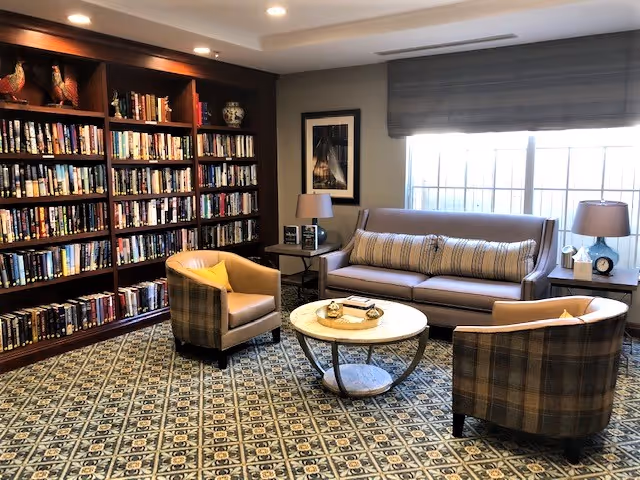 A cozy living room area with a large wooden bookshelf filled with books along one wall. The room has a patterned carpet, a round coffee table in the center, a gray sofa with striped cushions, and two plaid armchairs with yellow accent pillows. There are two side tables with lamps and a framed picture on the wall next to a large window with blinds.