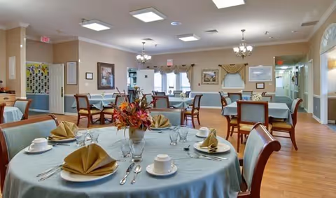 A dining room in a senior living facility with round tables covered in light blue tablecloths, each set with folded napkins, cups, saucers, glasses, and silverware. The room has wooden chairs, chandeliers, framed artwork on the walls, and large windows with curtains letting in natural light.