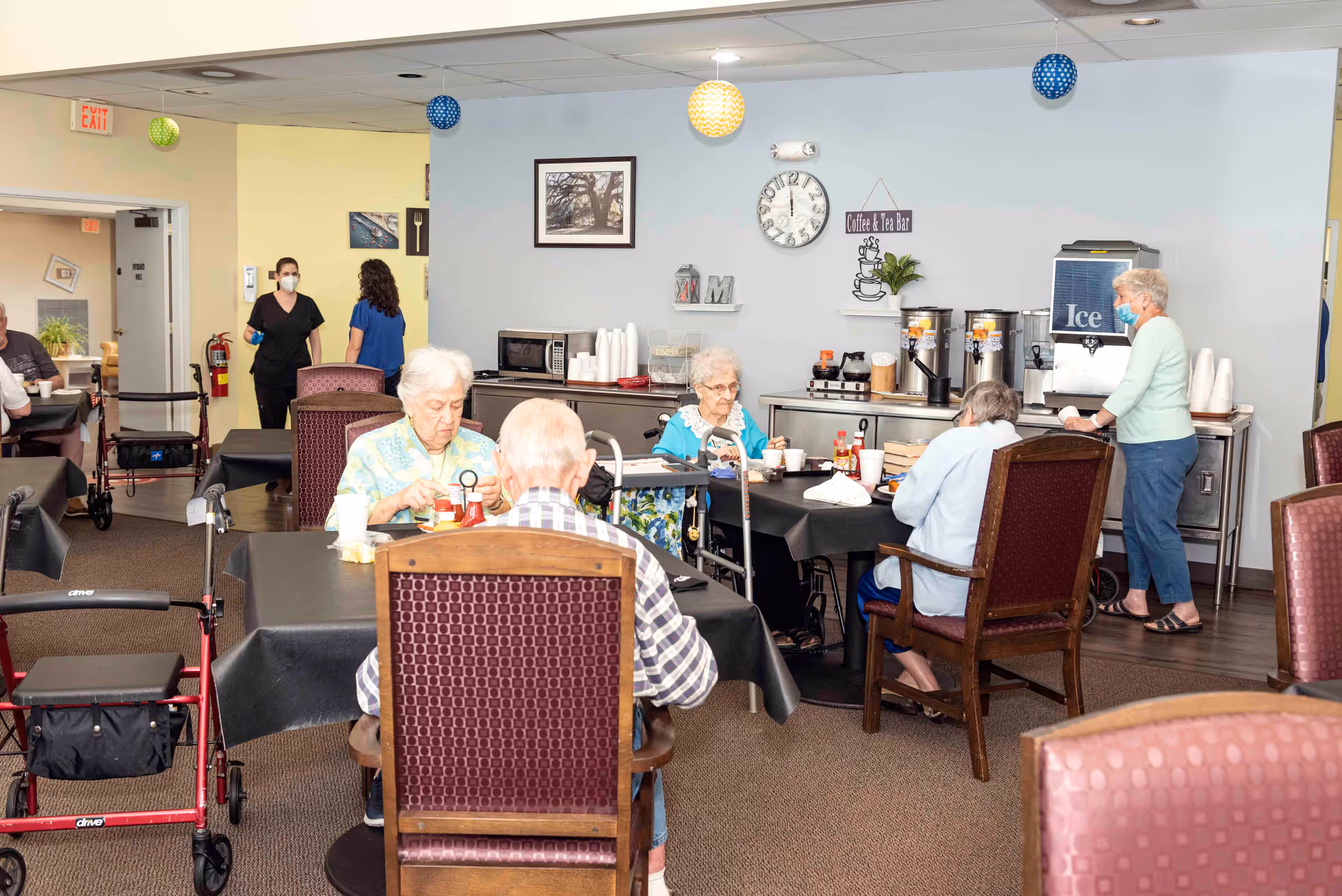 A dining area in a senior living facility with elderly residents seated at tables covered with black tablecloths, eating and drinking. A woman wearing a mask stands near a coffee and tea bar with dispensers and an ice machine. Two staff members, also wearing masks, are seen in the background near a hallway. The room is decorated with hanging paper lanterns and has framed pictures on the wall.