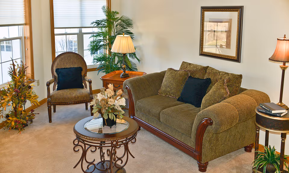 A cozy living room with a green upholstered sofa adorned with multiple cushions, a round glass-top coffee table with a floral arrangement, an armchair with a dark cushion, two side tables each with a lamp, a framed picture on the wall, and several plants near the windows.