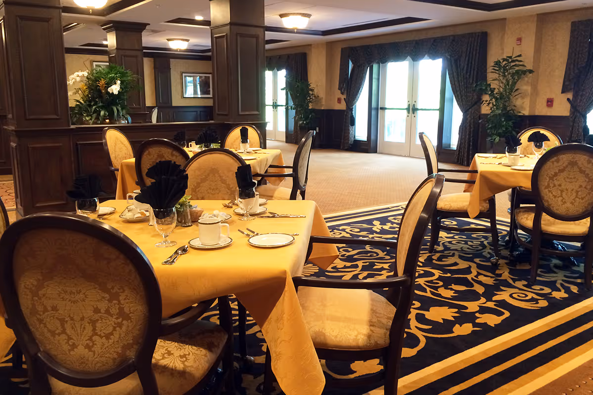 Elegant dining room with tables covered in yellow tablecloths, set with plates, cups, silverware, and black folded napkins. The room features upholstered chairs with patterned fabric, dark wood paneling, large windows with curtains, and decorative plants.