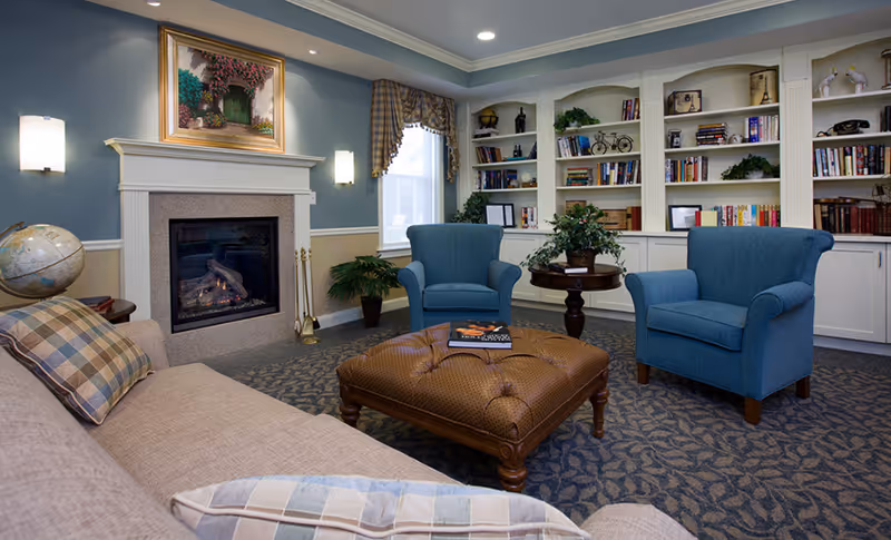 A cozy living room with a beige sofa adorned with plaid pillows, two blue armchairs, a large tufted ottoman with a book on top, a fireplace with a painting above it, built-in white bookshelves filled with books and decorative items, and a window with plaid curtains.
