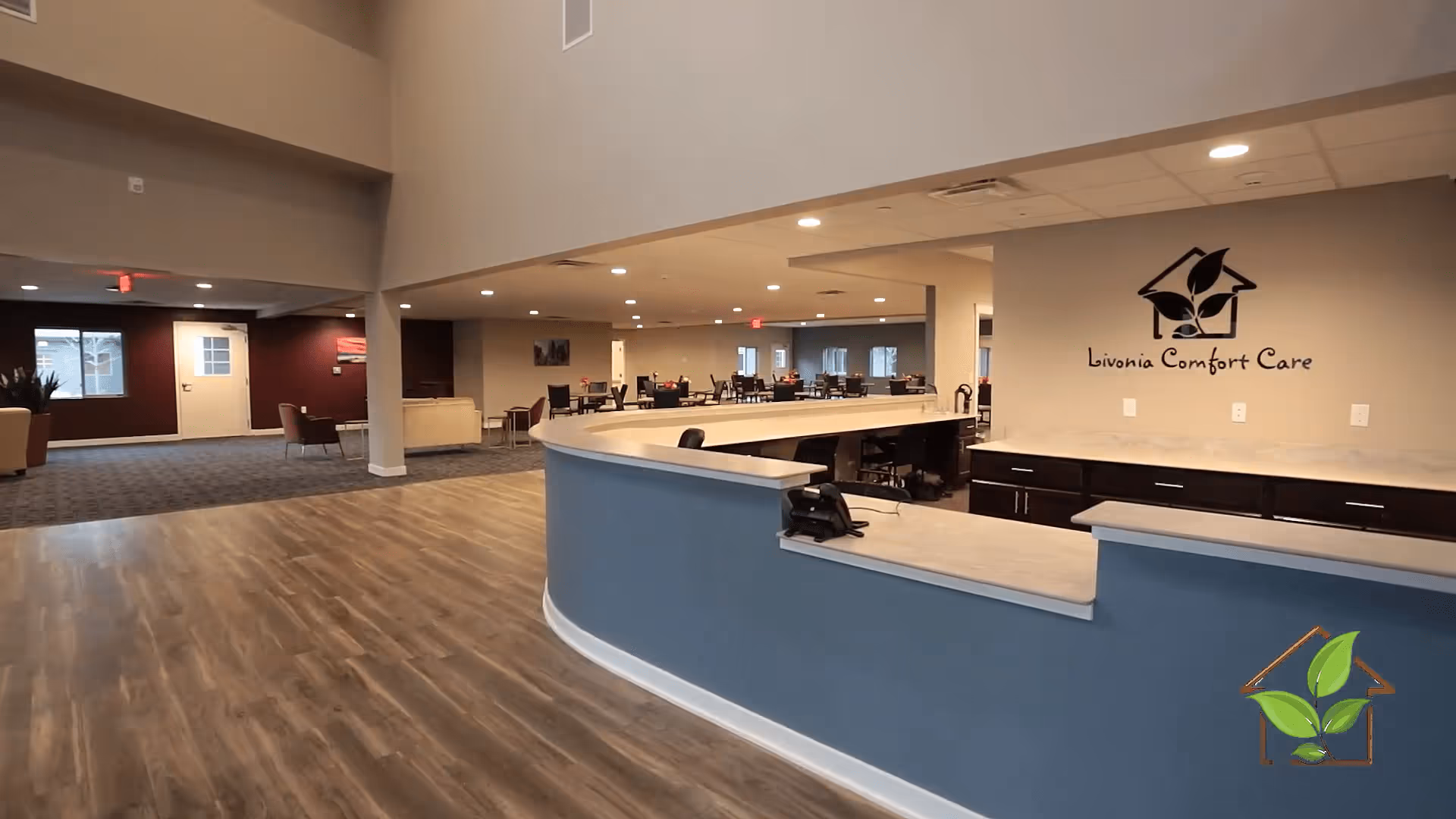 Interior view of Livonia Comfort Care facility showing a spacious reception area with a curved counter, wood flooring, and a seating area with chairs and tables in the background. The facility's logo and name are displayed on the wall behind the counter.