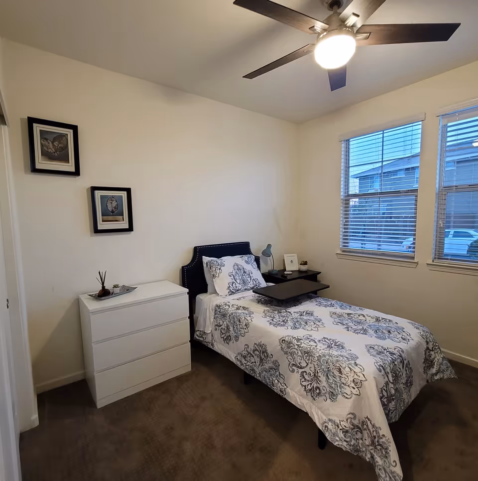 A small bedroom with a single bed covered in a white and blue patterned bedspread. There is a black headboard and a black overbed table on the bed. Next to the bed is a black nightstand with a small lamp, a framed photo, and a small plant. On the left side of the room is a white three-drawer dresser with decorative items on top. Two framed pictures hang on the wall above the dresser. The room has beige carpet, white walls, a ceiling fan with a light, and two windows with blinds showing an outside view of neighboring buildings and parked cars.