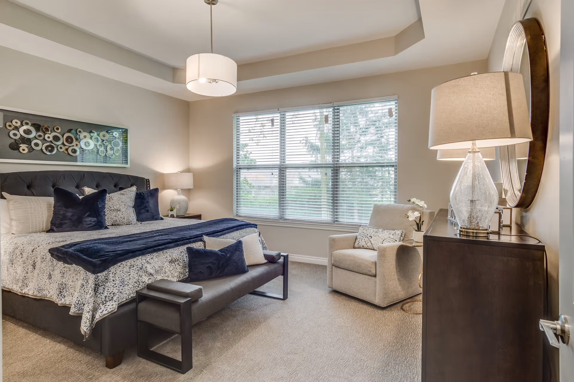 A well-lit bedroom with a large bed featuring a dark upholstered headboard, white and blue bedding, and multiple pillows. At the foot of the bed is a bench with a dark cushion and pillows. To the right, there is a beige armchair with a decorative pillow and a small round side table with a white flower pot. A dark wooden dresser with a glass lamp and a round mirror above it is positioned against the right wall. The room has a large window with blinds, beige walls, and a ceiling light fixture.