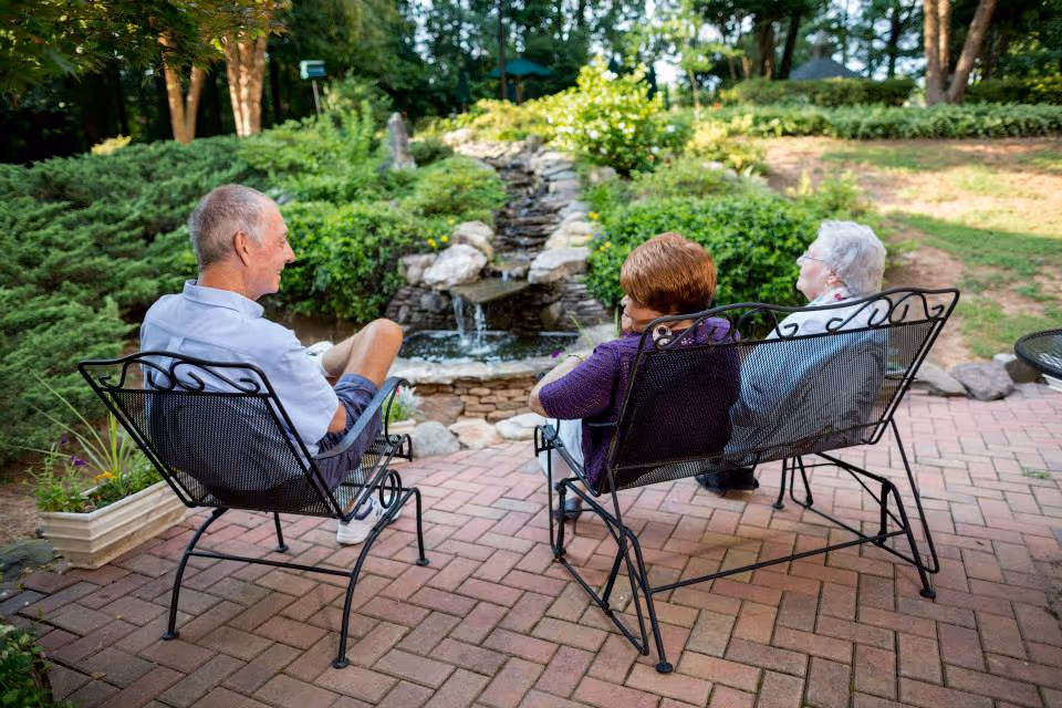 Three elderly people sitting on black metal outdoor chairs and bench on a brick patio, facing a small stone waterfall feature surrounded by greenery in a garden setting.