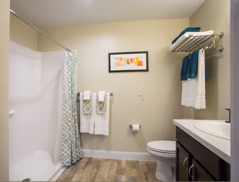 A clean and modern bathroom featuring a white bathtub with a blue and white patterned shower curtain, a toilet, a dark wood vanity with a white countertop and sink, and several neatly folded towels on a wall-mounted rack and towel bar. A colorful framed artwork hangs on the beige wall above the toilet paper holder.