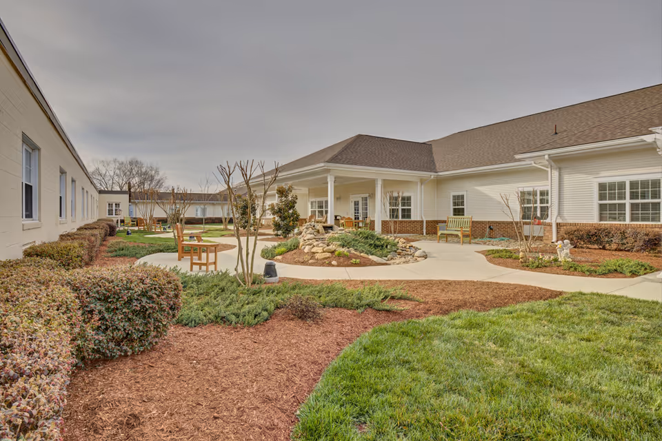 Outdoor courtyard area of a senior living facility with paved walkways, landscaped garden beds, bushes, small trees, and several wooden benches. The building surrounding the courtyard is single-story with white siding and multiple windows. The sky is overcast.