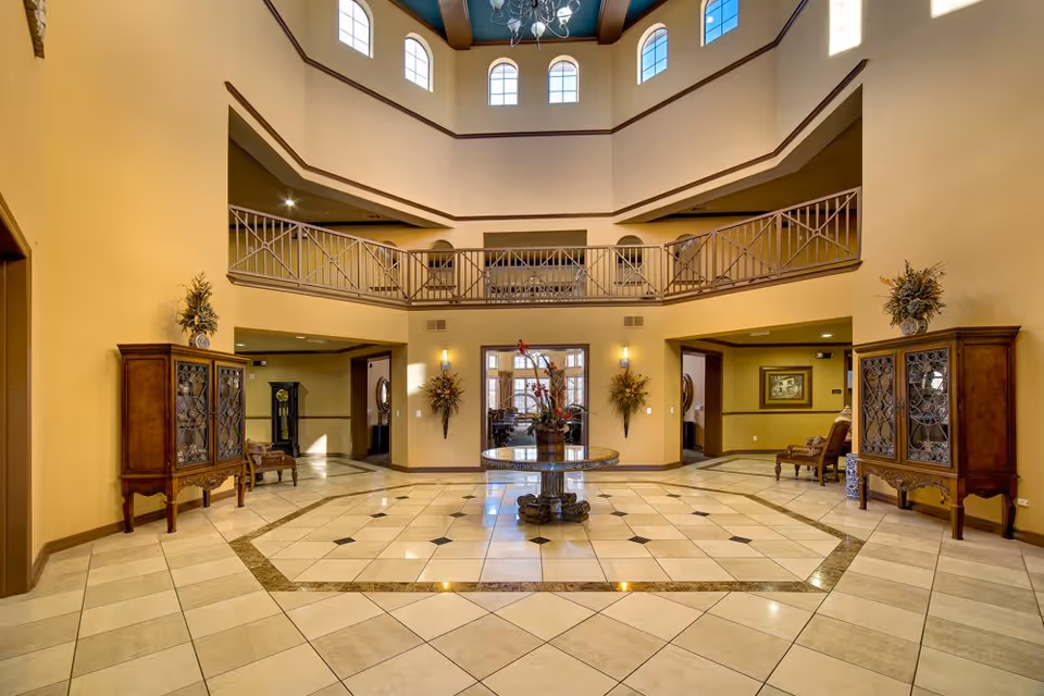 Spacious two-story lobby with a tiled floor, central round table with flowers, balcony railing and decorative cabinets and seating.