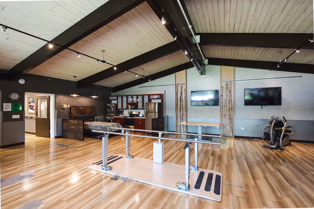 Spacious senior living rehab/interior room with parallel walking bars in the center, exercise bike, bed, kitchenette and wall-mounted TVs under exposed beams.