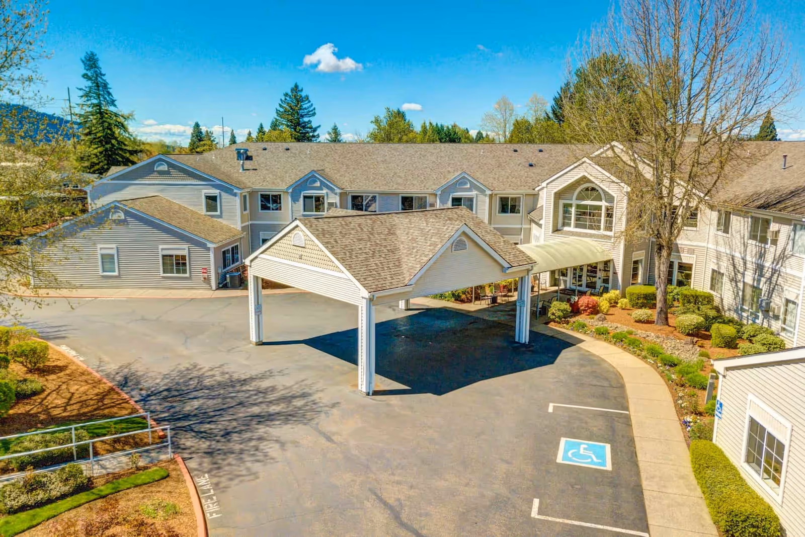 Exterior view of The Oaks at Lebanon senior living facility showing a large two-story building with a covered entrance driveway, landscaped bushes and trees, a clear blue sky, and a handicapped parking space in front.
