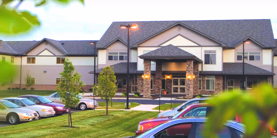 Front exterior view of Mankato Lodge Senior Living building with a covered entrance supported by stone pillars, surrounded by a parking lot with several cars and small trees on a grassy lawn.