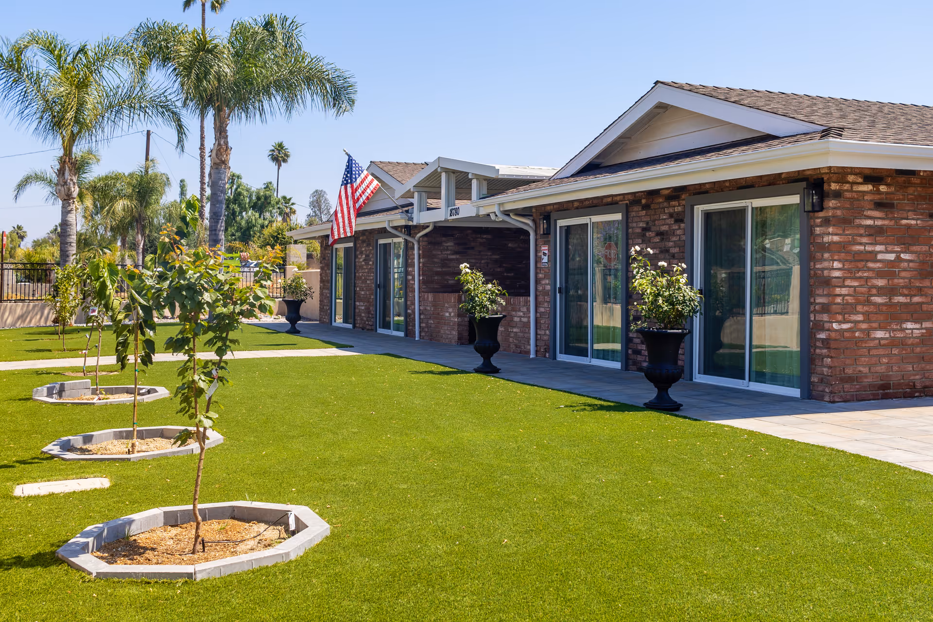 Exterior view of a single-story brick building with sliding glass doors, potted plants, and an American flag. The building is surrounded by a well-maintained lawn with small trees planted in circular beds and palm trees in the background under a clear blue sky.