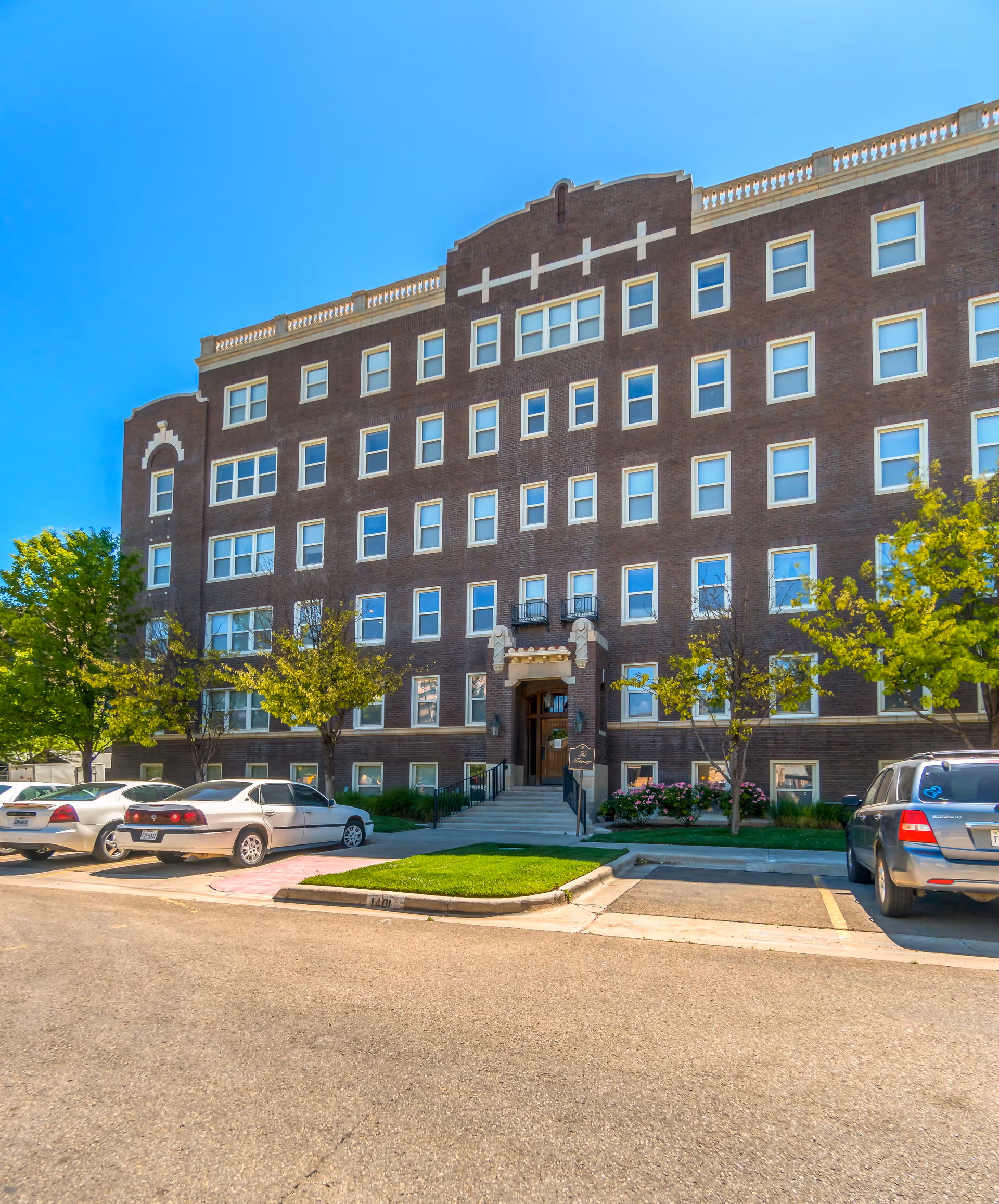 Exterior view of a multi-story brick building with numerous windows, a central entrance with steps and decorative elements, surrounded by trees and parked cars under a clear blue sky.