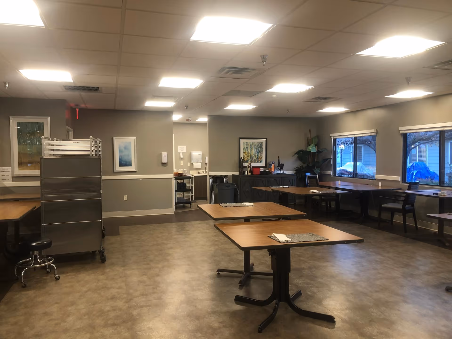 A spacious dining room with several wooden tables and chairs arranged neatly. The room has large windows on one side allowing natural light to enter. The walls are painted gray and decorated with framed artwork. There is a metal rack on wheels and a small counter area with various items in the background.