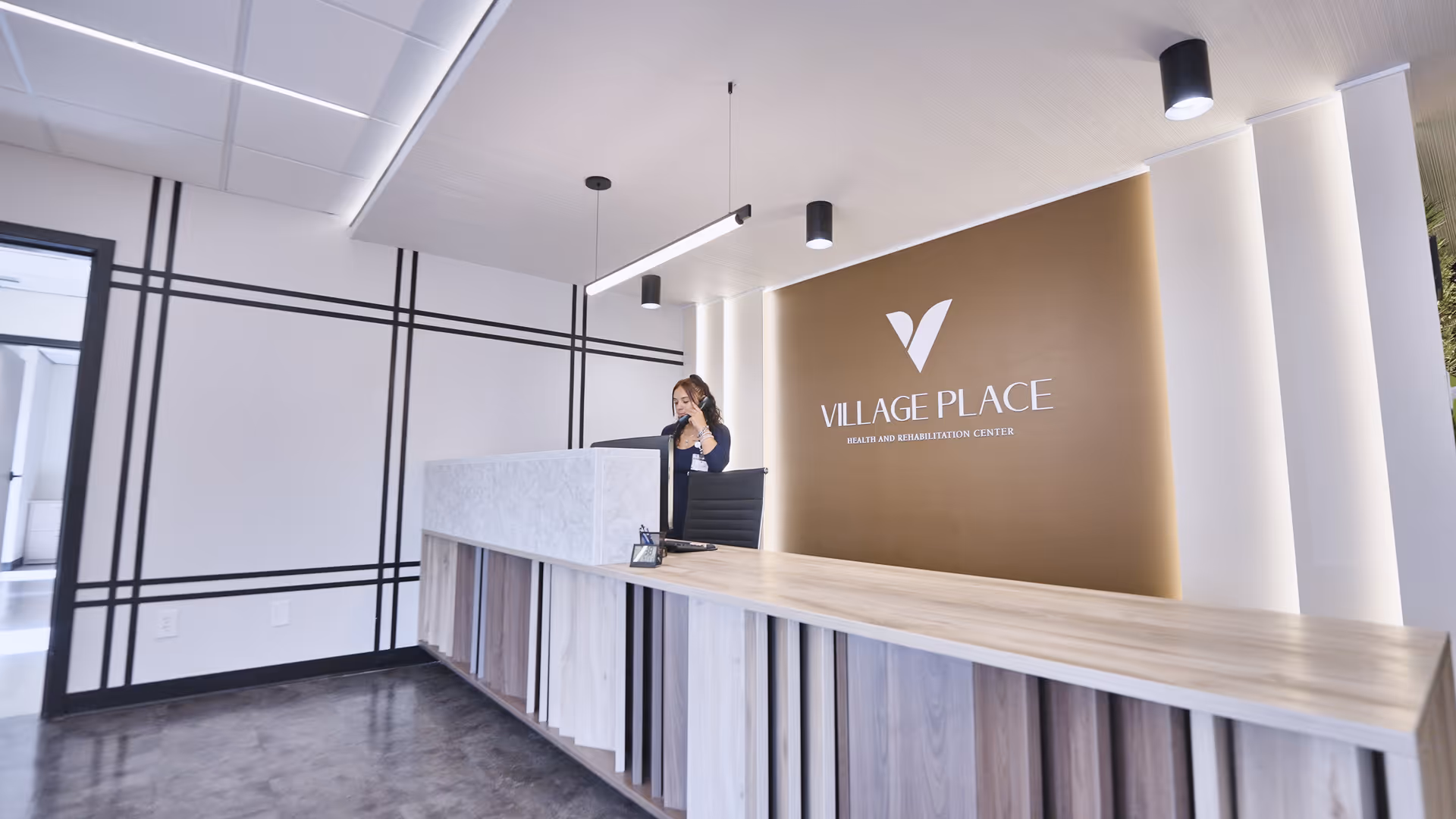 Reception area of Village Place Health and Rehabilitation Center with a modern wooden reception desk and a woman standing behind it talking on the phone. The background features a brown wall with the facility's logo and name, and the room has white walls with black geometric lines and ceiling lights.