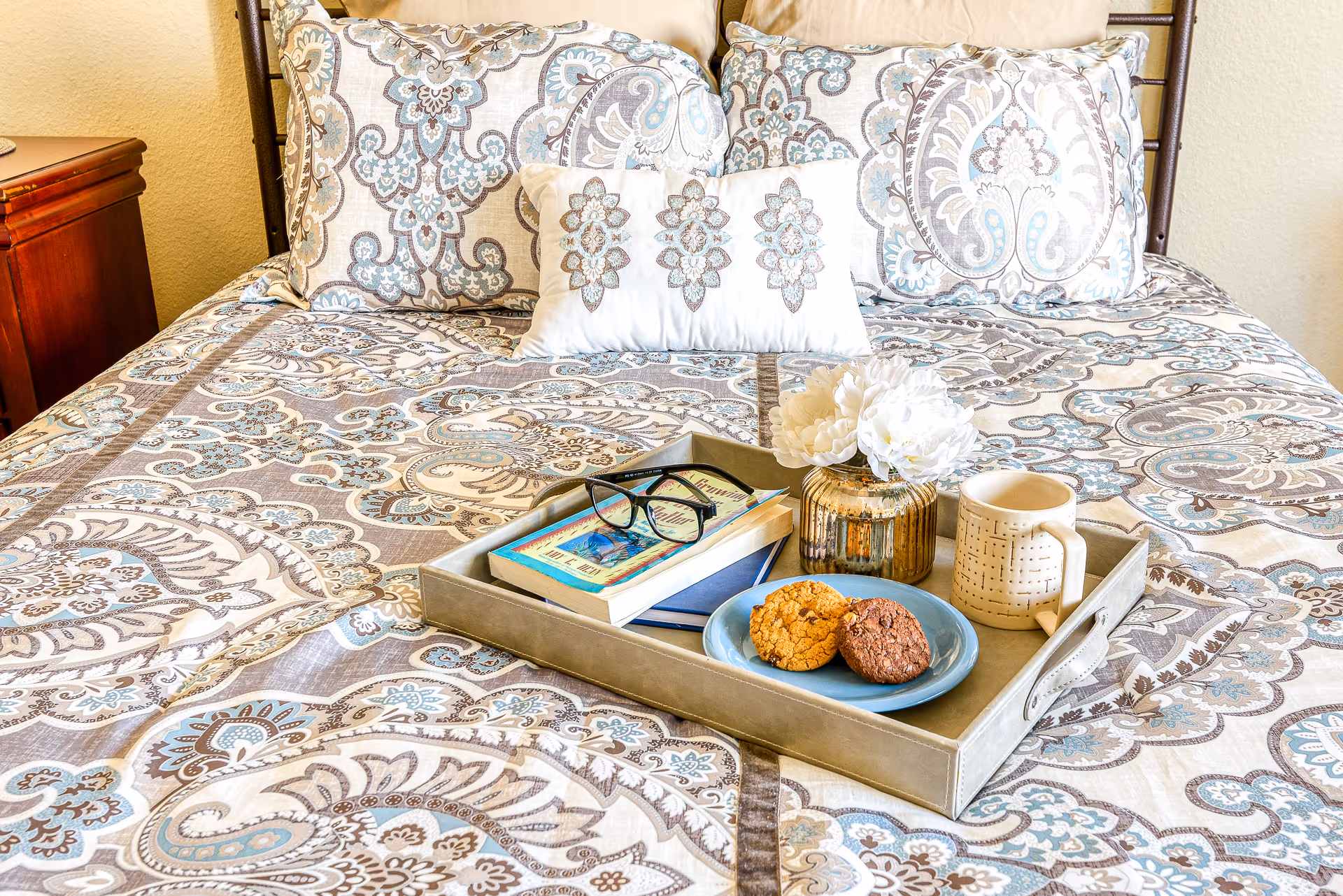 A decorative tray holding cookies, a mug, books, glasses, and a small vase sits on a patterned bed with pillows in a bedroom.
