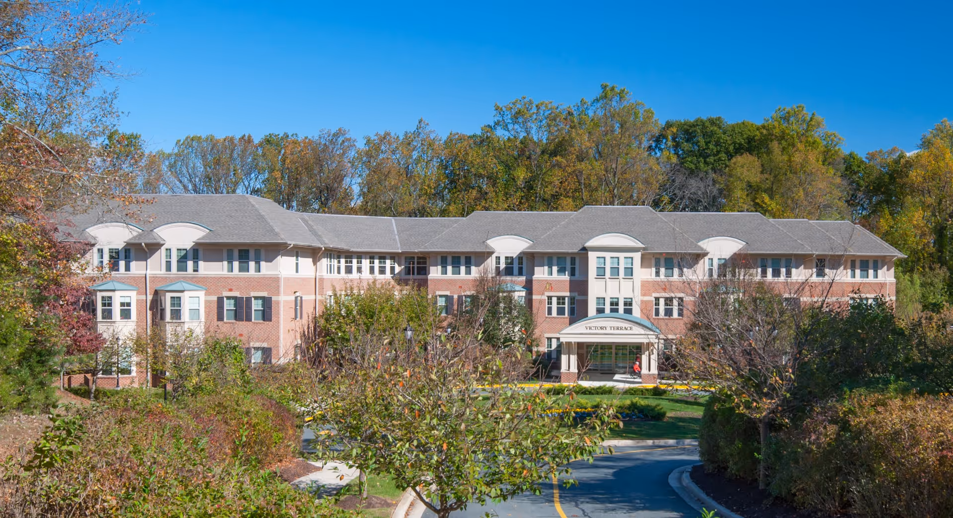 Exterior view of Victory Terrace, a three-story senior living facility surrounded by trees and landscaped bushes under a clear blue sky.