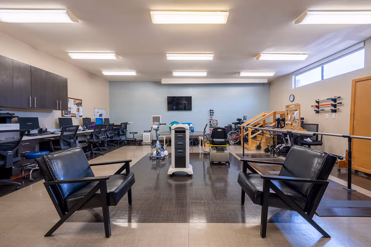 A spacious rehabilitation or therapy room with two black leather chairs in the foreground, exercise and therapy equipment in the center, and a row of desks with computers and chairs along the left wall. The room has bright overhead lighting, a wall-mounted TV, and large windows near the ceiling on the right side.