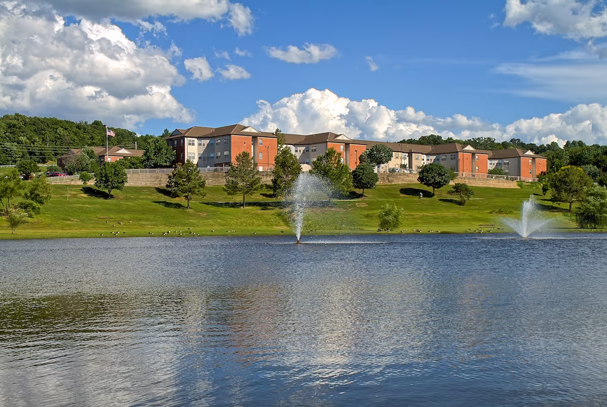Lakeside view of a multi-story senior living building on a grassy hill with two fountain jets in the pond under a partly cloudy sky.