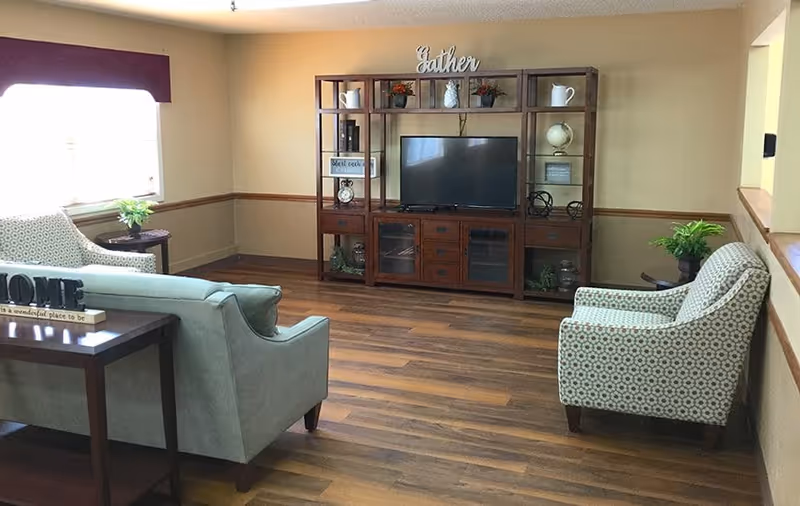 A cozy living room with wooden flooring, a large wooden entertainment center holding a flat-screen TV and decorative items, two patterned armchairs, a light blue sofa, and a side table with a plant. The walls are painted beige with a wooden chair rail, and a window with a dark valance lets in natural light.