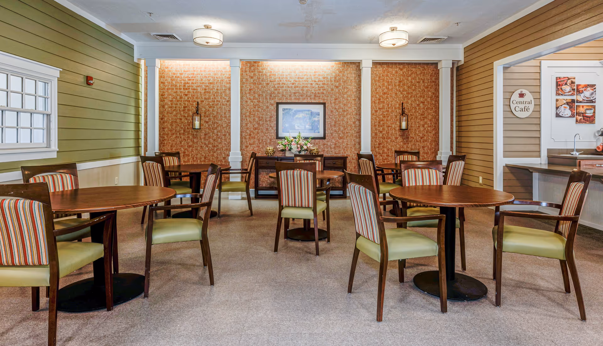 A cozy dining area with several round wooden tables and chairs featuring green seats and striped backs. The walls are decorated with green and brown paneling, a framed picture, and a sign that reads 'Central Café' near a small counter with a sink. The room is softly lit by ceiling lights and wall sconces.
