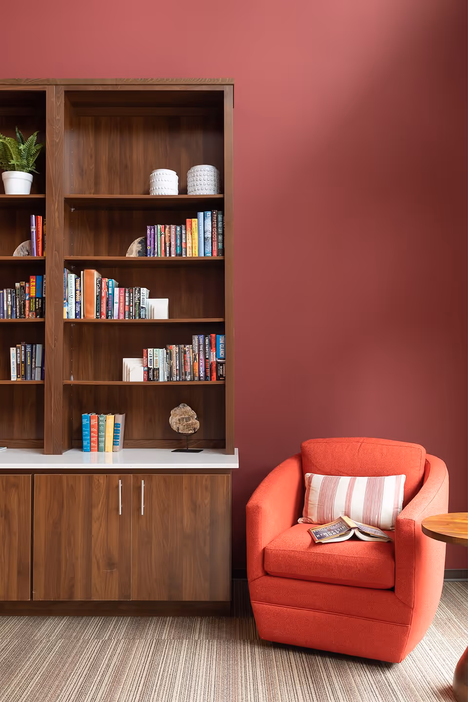 Cozy reading nook with a red upholstered armchair featuring a striped pillow and an open book, next to a wooden bookshelf filled with books and decorative items against a red wall.