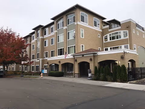 Exterior view of a multi-story senior living facility building with beige siding, multiple windows, an American flag near the entrance, and a tree with red leaves on the left side.