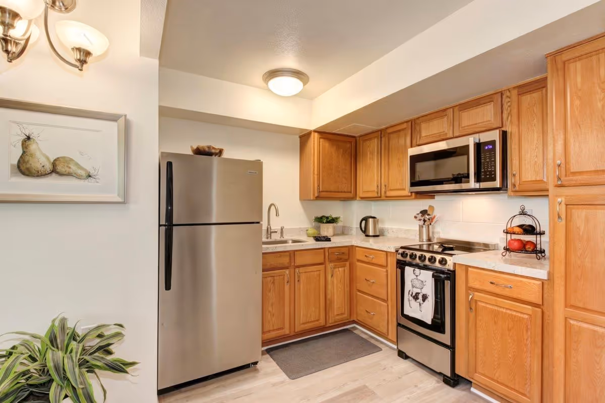 A modern kitchen with wooden cabinets, a stainless steel refrigerator, a stainless steel microwave above a stove, a sink with a faucet, a kettle, a fruit stand with apples and oranges, a small plant on the counter, and a framed picture of pears on the wall. The floor is light-colored wood, and there is a gray mat in front of the sink.