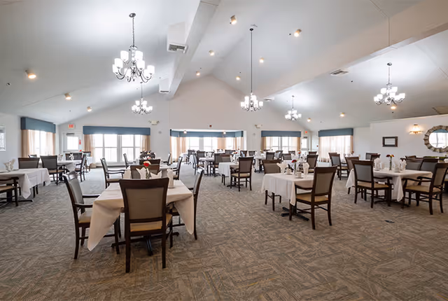 A spacious dining room with multiple tables covered in white tablecloths, each set with chairs and small floral centerpieces. The room features large windows with blue valances, carpeted floors, and several elegant chandeliers hanging from a high ceiling.