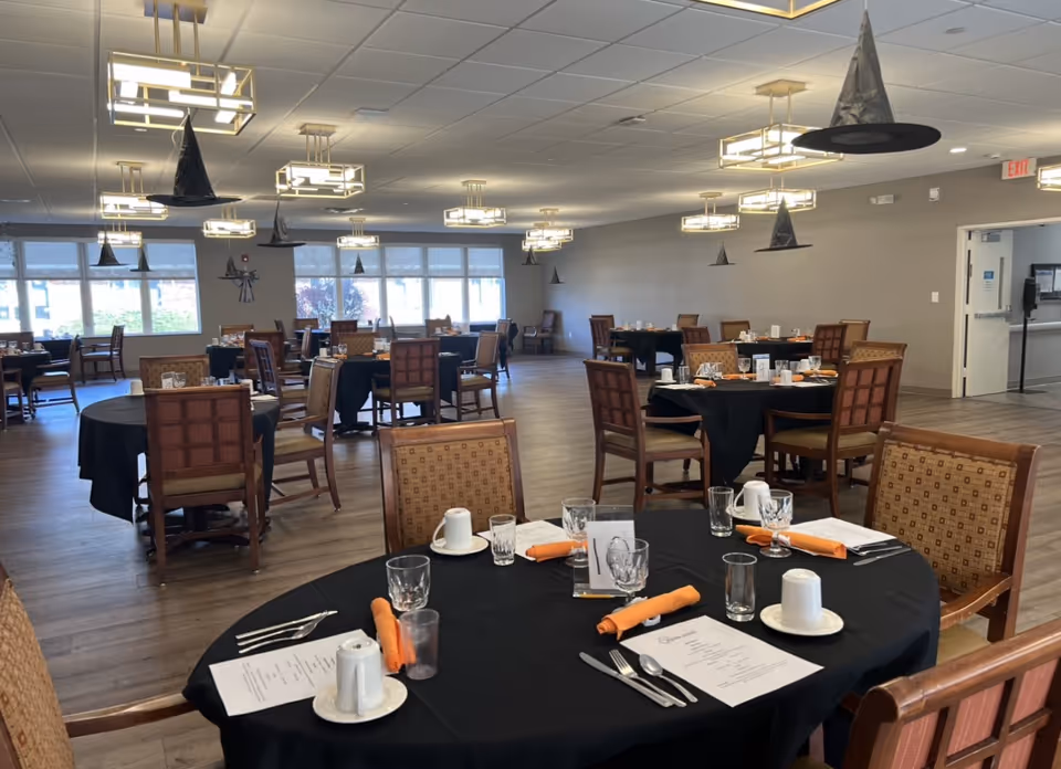A spacious dining room with multiple round tables covered with black tablecloths, each set with menus, glasses, cups, silverware, and orange napkins. The ceiling has modern light fixtures and hanging black witch hats as decorations. Large windows allow natural light to brighten the room.