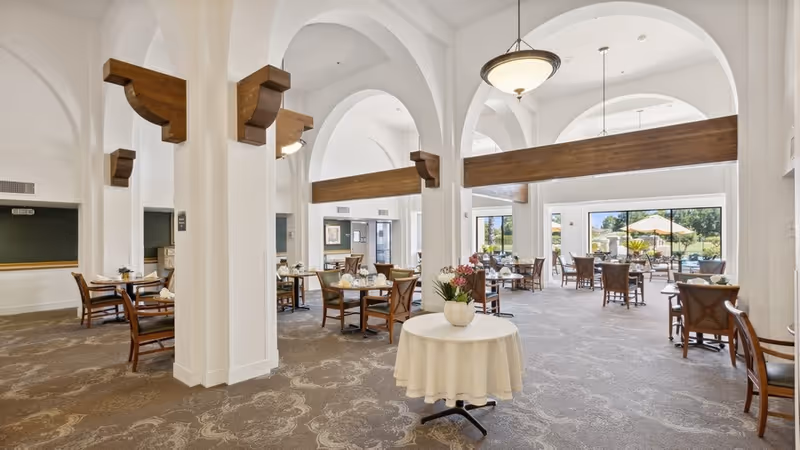 Spacious dining area with multiple wooden tables and chairs arranged neatly. The room features high white arched ceilings with wooden beams, large windows letting in natural light, and a round table with a white tablecloth and a flower vase in the center.