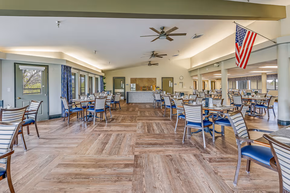 Spacious dining room with multiple tables and chairs arranged neatly. The room features large windows on the left side allowing natural light to enter, ceiling fans, and an American flag hanging from the ceiling on the right. The floor has a wood-like pattern, and there is a counter area at the far end of the room.