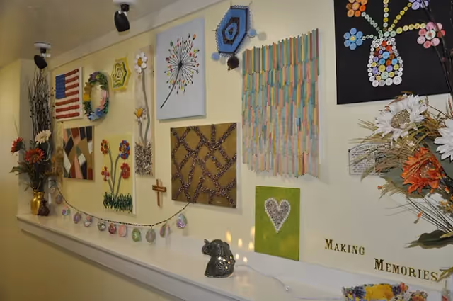 A hallway wall decorated with various colorful handmade art pieces including flowers, an American flag, a heart, and abstract designs. There are also flower arrangements on both ends of the shelf below the artwork. The words 'Making Memories' are displayed on the wall.