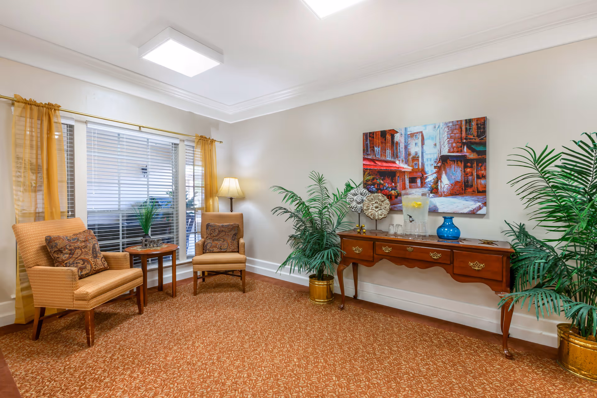 A cozy sitting area with two upholstered armchairs and a small round wooden table between them. The chairs have patterned cushions. Behind the chairs is a large window with white blinds and sheer yellow curtains. To the right, there is a wooden console table with decorative items including a blue vase, a glass pitcher with lemon water, and some glasses. Two large potted plants flank the console table. A colorful painting of a street scene hangs above the console. The room has a patterned carpet and soft lighting from a floor lamp in the corner.