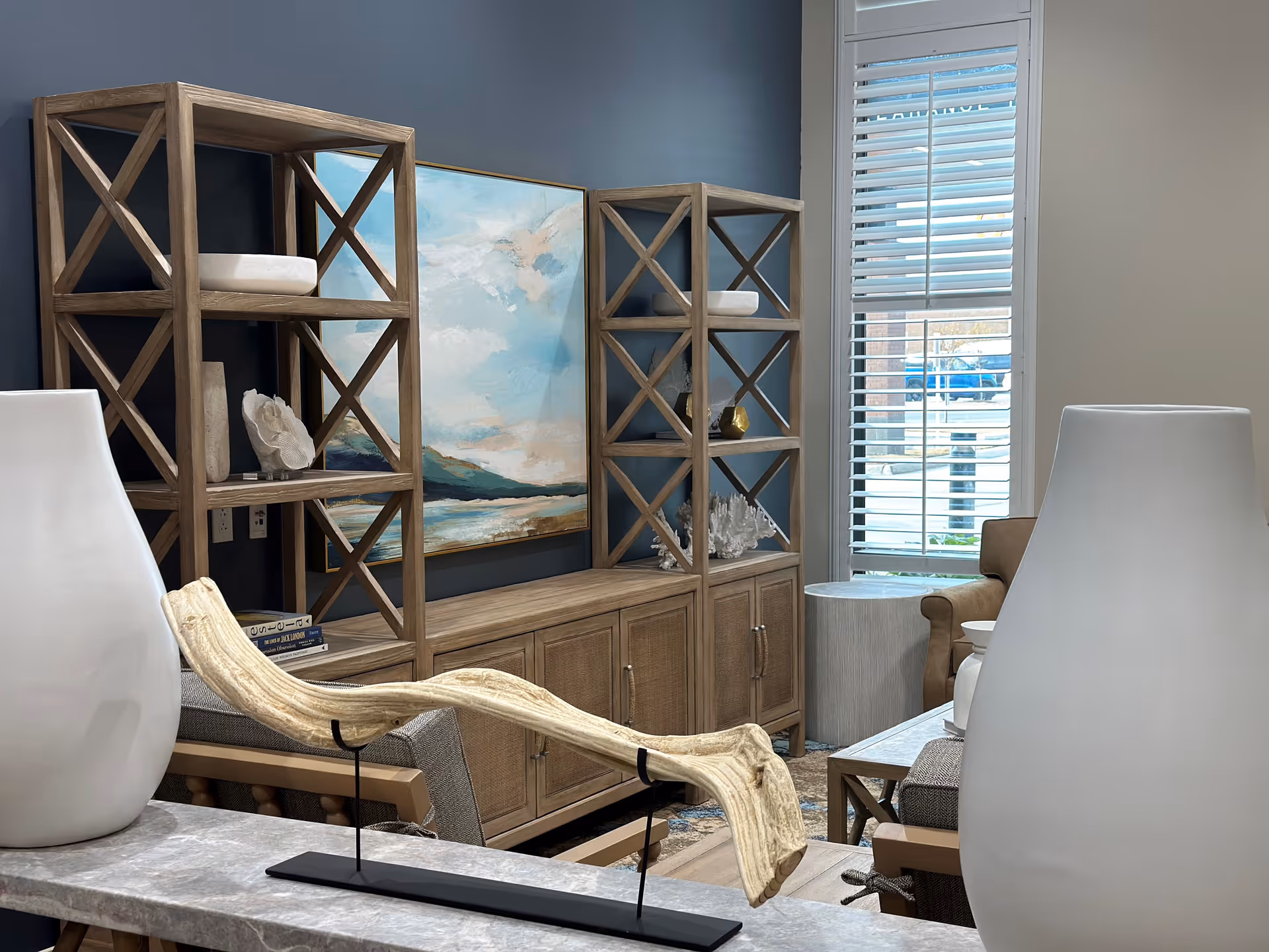Communal living room with wooden display shelves, a framed seascape painting, seating, and decorative accents by a window with blinds.