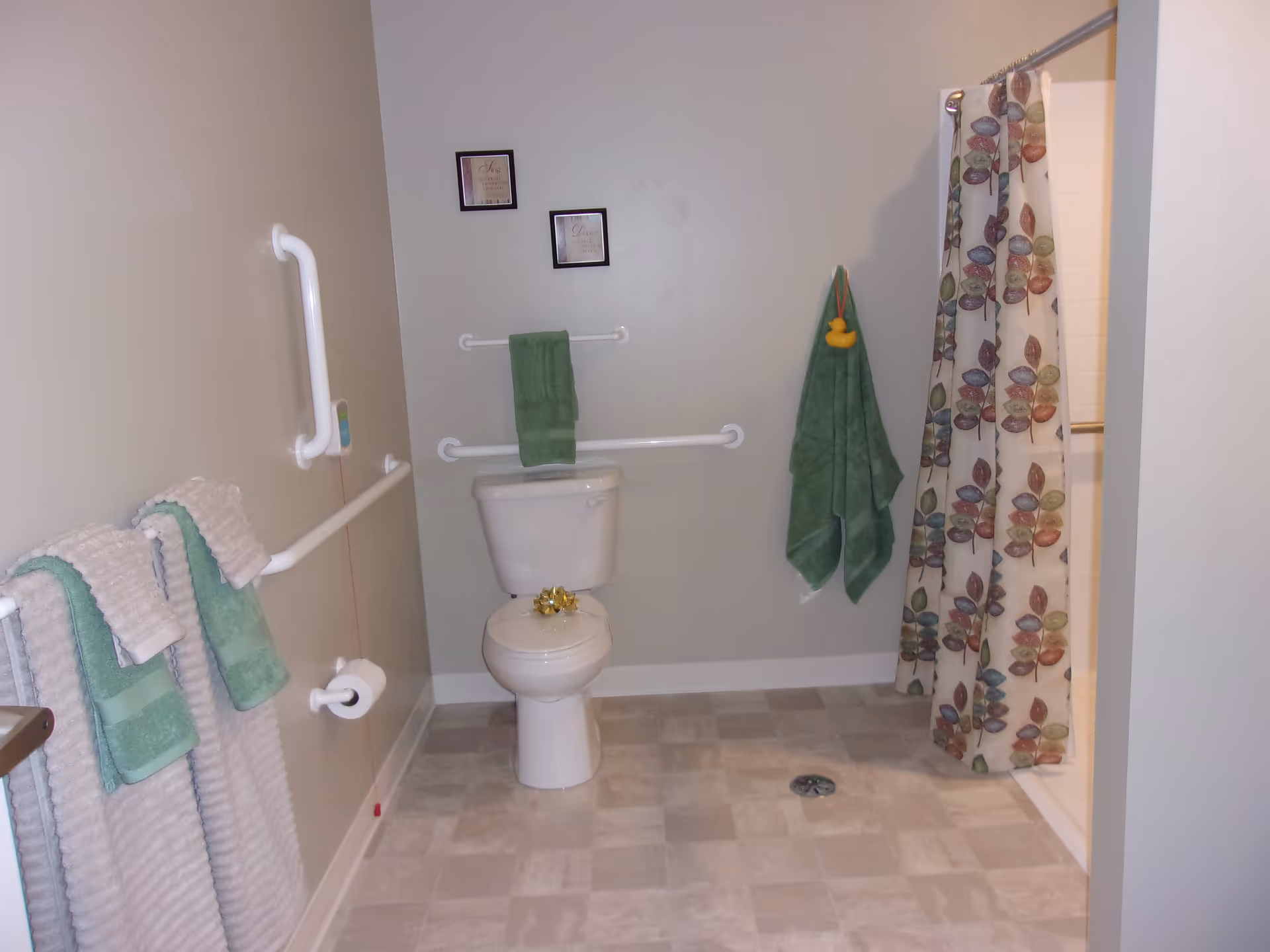 A bathroom with a toilet in the center, grab bars on the walls, green towels hanging on towel racks and hooks, a shower with a colorful leaf-patterned curtain on the right, and a tiled floor.