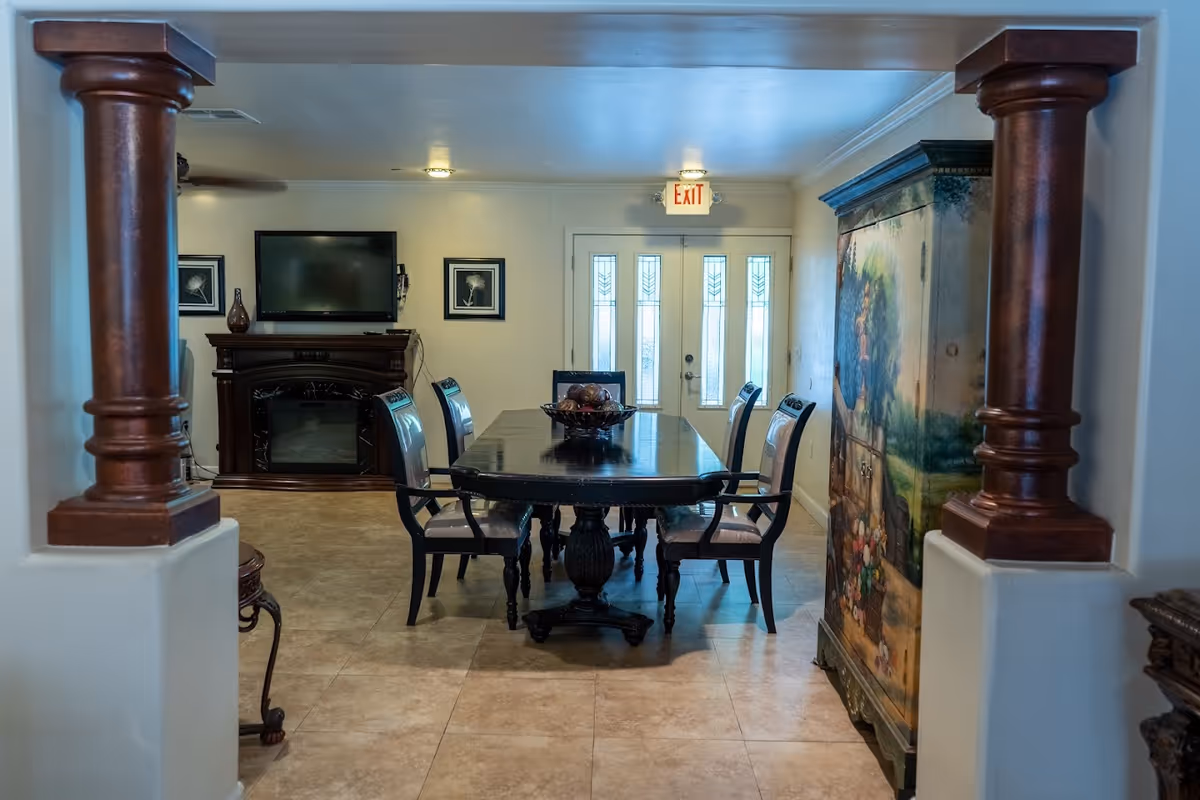 Interior view of a dining room with a dark wooden dining table and six chairs. The room features tiled flooring, two large wooden columns framing the entrance, a decorative cabinet with a painted floral scene on the right, a wall-mounted TV above a fireplace on the left, and a set of glass-paneled double doors with an exit sign above them at the back.