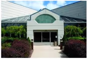 Entrance to a building with glass double doors, surrounded by greenery including bushes and small trees, with a glass roof structure above the entrance.