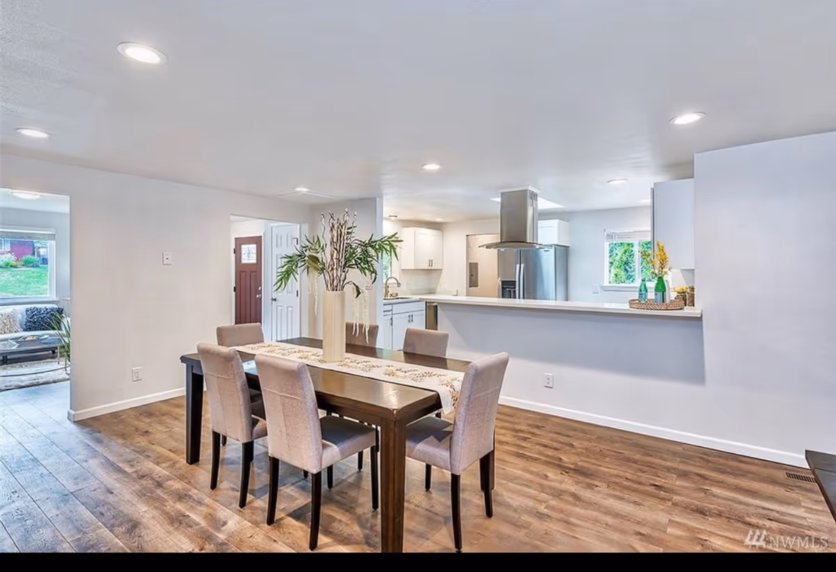 Open-plan dining area with a wooden table and upholstered chairs facing a white kitchen with stainless-steel appliances.