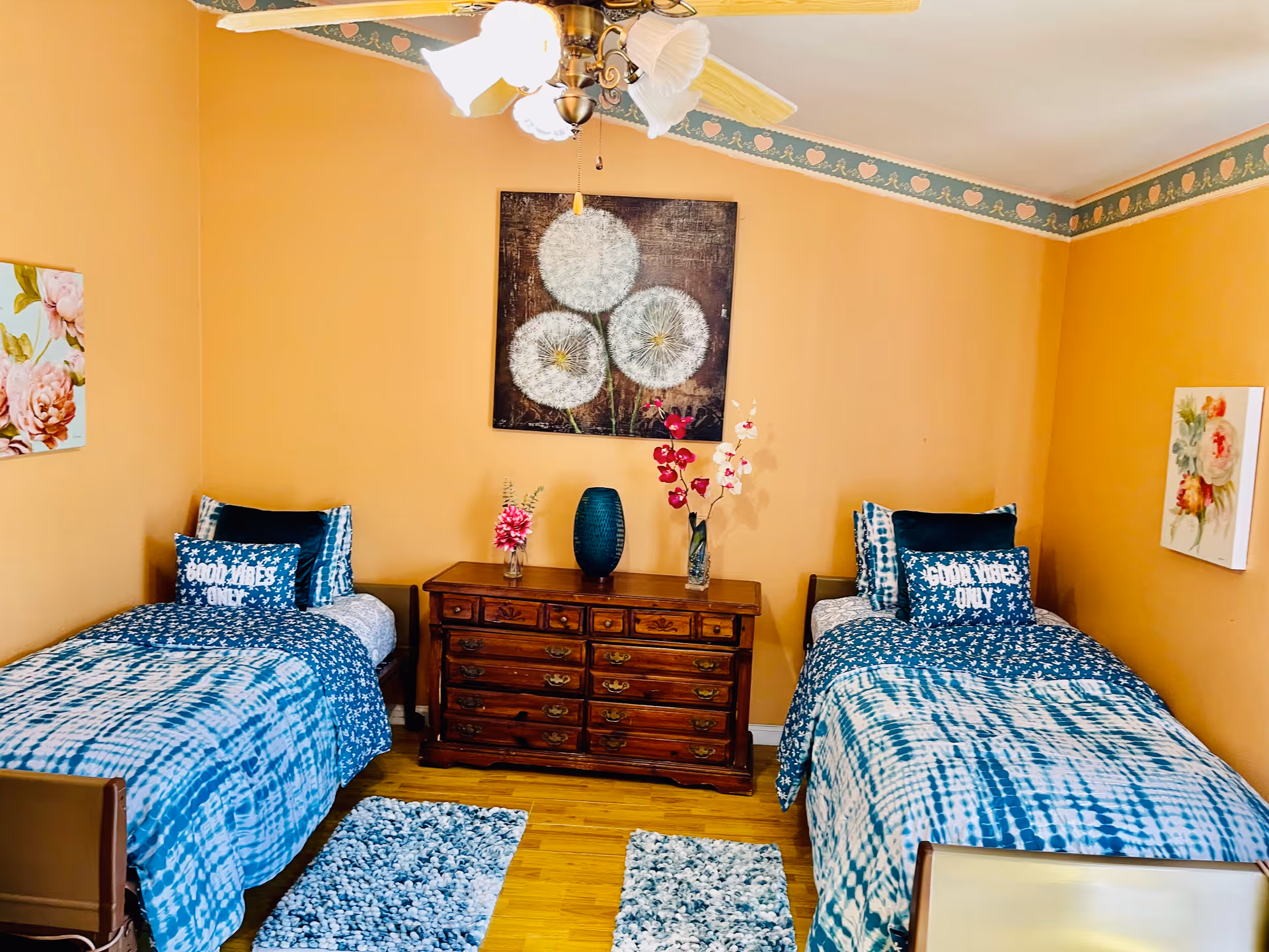A cozy bedroom with two twin beds featuring blue and white patterned bedding and pillows that say 'GOOD VIBES ONLY'. Between the beds is a wooden dresser with decorative vases and flowers. The walls are painted a warm beige color with floral artwork and a ceiling fan with lights above.