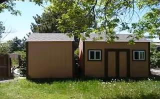 Two small brown storage sheds on a grassy lawn with trees and a wooden fence in the background under a clear sky.